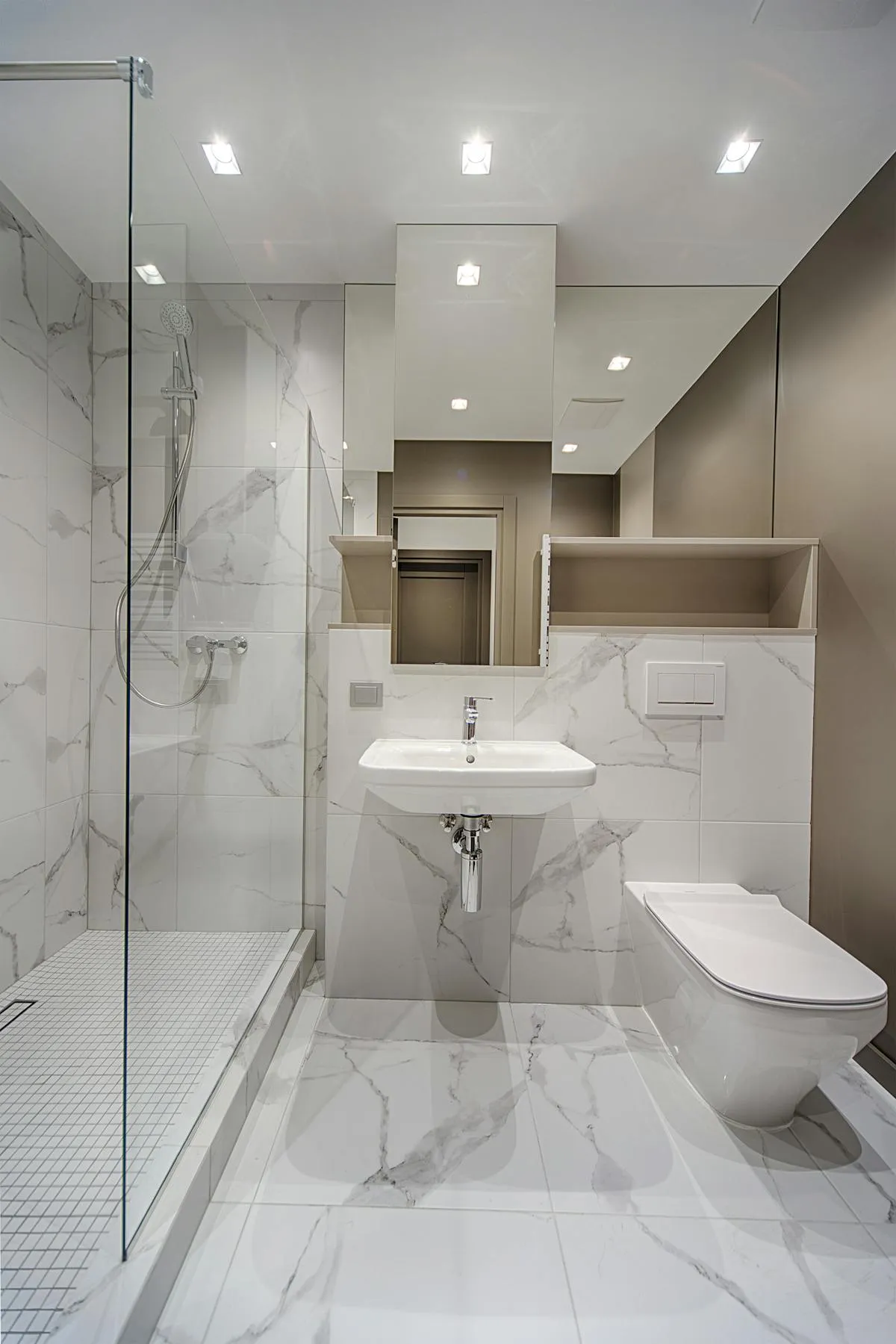 A bright marble bathroom interior with a wall-hung toilet, glass shower, and recessed lighting